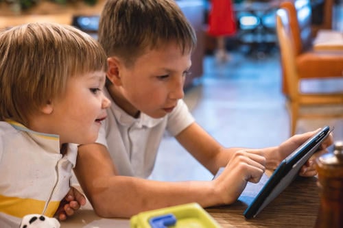 Boy and toddler using tablet online on internet while waiting orders in restaurant. 