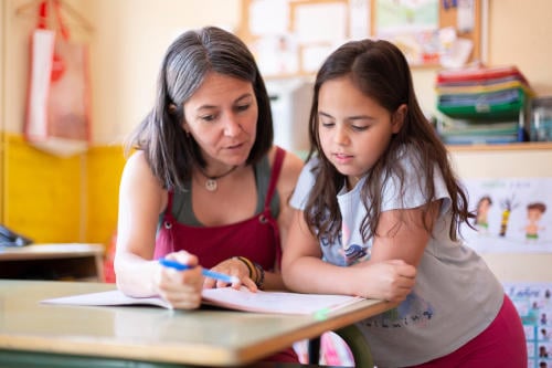 Young adult female teacher helping a primary school pupil with dyslexia in class to learn a lesson.
