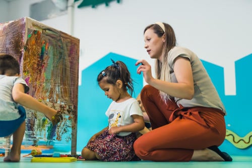 Cling film painting. Toddler painting with a sponge, brushes and paints on a cling film wrapped all the way round the wooden shelf unit. A teacher helping them. Creative activity for kids sensory skills development at the nursery school